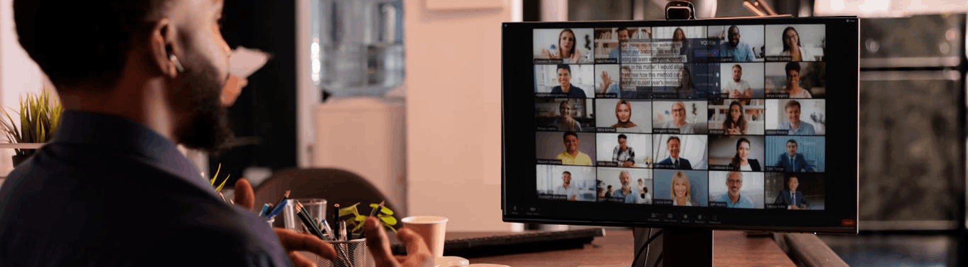Over-the-shoulder view of a man at a desk facing a large monitor showing a video-call grid with a teleprompter overlay